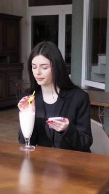 Bir kız sokakta masada otururken akıllı telefon kullanıyor ve kokteyl içiyor..