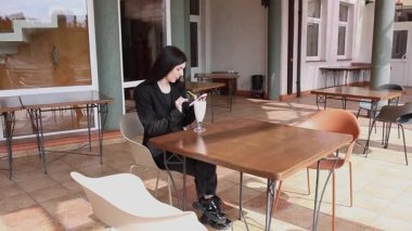 Bir kız sokakta masada otururken akıllı telefon kullanıyor ve kokteyl içiyor..