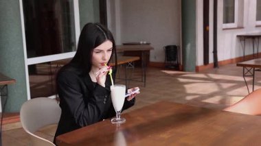 Bir kız sokakta masada otururken akıllı telefon kullanıyor ve kokteyl içiyor..