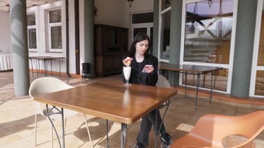 Bir kız sokakta masada otururken akıllı telefon kullanıyor ve kokteyl içiyor..