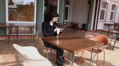 Bir kız sokakta masada otururken akıllı telefon kullanıyor ve kokteyl içiyor..