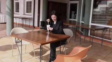 Bir kız sokakta masada otururken akıllı telefon kullanıyor ve kokteyl içiyor..