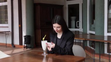 Bir kız sokakta masada otururken akıllı telefon kullanıyor ve kokteyl içiyor..