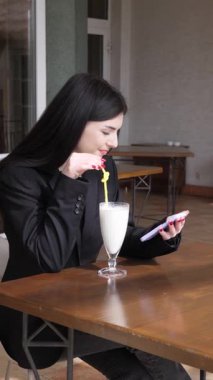 Bir kız sokakta masada otururken akıllı telefon kullanıyor ve kokteyl içiyor..