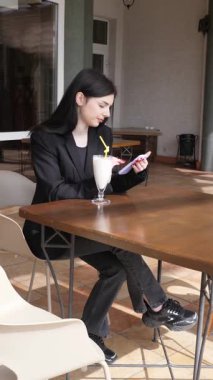 Bir kız sokakta masada otururken akıllı telefon kullanıyor ve kokteyl içiyor..