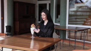 Bir kız sokakta masada otururken akıllı telefon kullanıyor ve kokteyl içiyor..