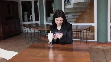 Bir kız sokakta masada otururken akıllı telefon kullanıyor ve kokteyl içiyor..