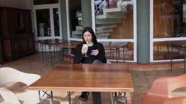 Bir kız sokakta masada otururken akıllı telefon kullanıyor ve kokteyl içiyor..