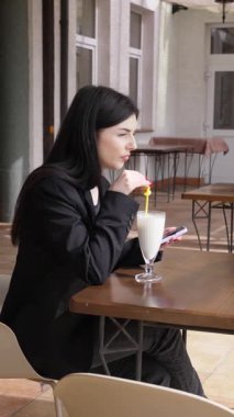 Bir kız sokakta masada otururken akıllı telefon kullanıyor ve kokteyl içiyor..