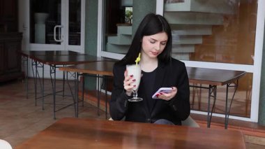 Bir kız sokakta masada otururken akıllı telefon kullanıyor ve kokteyl içiyor..