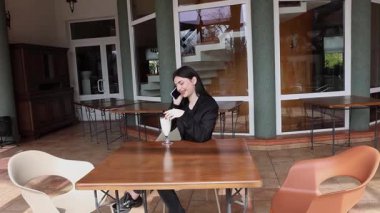 Bir kız sokakta masada otururken akıllı telefon kullanıyor ve kokteyl içiyor..