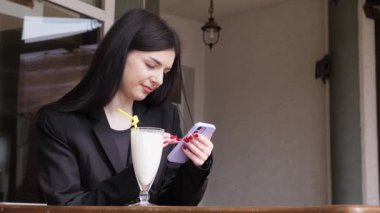 Bir kız sokakta masada otururken akıllı telefon kullanıyor ve kokteyl içiyor..