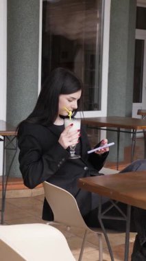 Bir kız sokakta masada otururken akıllı telefon kullanıyor ve kokteyl içiyor..