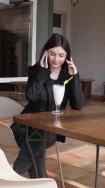 Bir kız sokakta masada otururken akıllı telefon kullanıyor ve kokteyl içiyor..