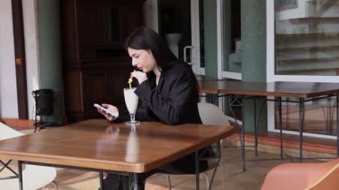 Bir kız sokakta masada otururken akıllı telefon kullanıyor ve kokteyl içiyor..
