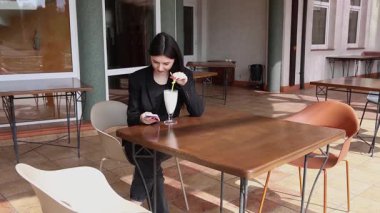 Bir kız sokakta masada otururken akıllı telefon kullanıyor ve kokteyl içiyor..