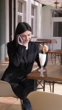 Bir kız sokakta masada otururken akıllı telefon kullanıyor ve kokteyl içiyor..