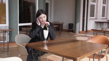 Bir kız sokakta masada otururken akıllı telefon kullanıyor ve kokteyl içiyor..