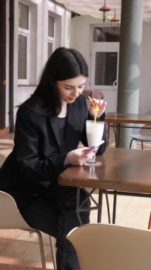 Bir kız sokakta masada otururken akıllı telefon kullanıyor ve kokteyl içiyor..