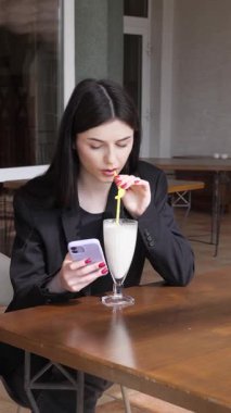 Bir kız sokakta masada otururken akıllı telefon kullanıyor ve kokteyl içiyor..
