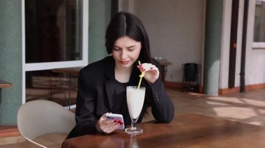 Bir kız sokakta masada otururken akıllı telefon kullanıyor ve kokteyl içiyor..