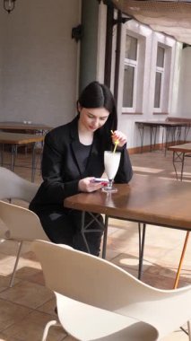 Bir kız sokakta masada otururken akıllı telefon kullanıyor ve kokteyl içiyor..