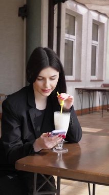 Bir kız sokakta masada otururken akıllı telefon kullanıyor ve kokteyl içiyor..