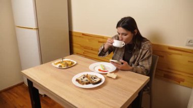 Güzel bir kız masada oturuyor, ev yapımı yemekler yiyor ve akıllı telefon kullanıyor..