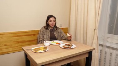 Güzel bir kız masada oturuyor, ev yapımı yemekler yiyor ve akıllı telefon kullanıyor..