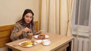 Bir kız masada oturuyor, akıllı telefona bakıyor ve çörekle kahve içiyor..