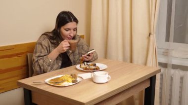 Bir kız masada oturuyor, akıllı telefona bakıyor ve çörekle kahve içiyor..