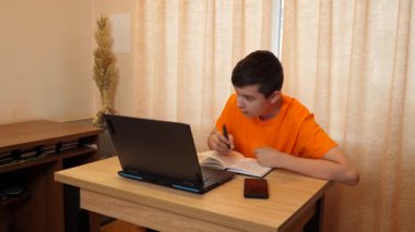 Bir genç, bir odada masada otururken dizüstü bilgisayarda ders çalışır..