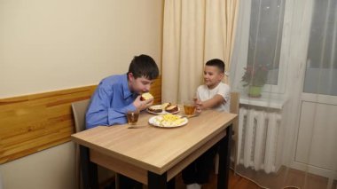 Çocuklar masada otururken sandviç, meyve yiyip çay içiyorlar.