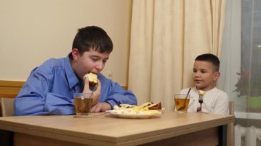 Çocuklar masada otururken sandviç, meyve yiyip çay içiyorlar.