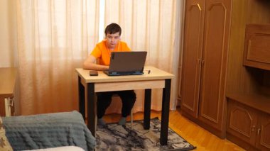 Bir genç, bir odada masada otururken dizüstü bilgisayarda ders çalışır..