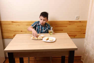 Bir çocuk masada otururken tatlı çörekler yer ve çay içer..