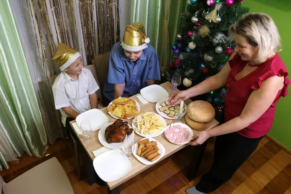 Evde şenlikli bir akşam yemeği: bir kadın geleneksel tabaklarla bir masa hazırlıyor, yeni yıl şapkalı çocuklar oyuncaklarla bir Noel ağacının yanında oturuyor..
