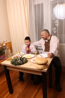 İşlemeli gömlekler içindeki baba ve oğul Noel 'i geleneksel yemek, ekmek ve Noel süslemeleriyle bir festival masasında kutluyorlar..