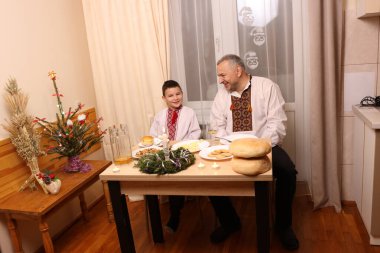 İşlemeli gömlekler içindeki baba ve oğul Noel 'i geleneksel yemek, ekmek ve Noel süslemeleriyle bir festival masasında kutluyorlar..