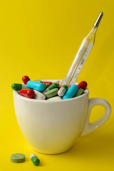 Renkli farklı haplar ve sarı arka plandaki bir bardağın içindeki tıbbi termometre.