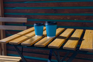 İki adet tek kullanımlık kahve fincanı, sakin bir ortamda ahşap ve turkuaz gölgelerin yer aldığı, rahat bir oturma alanındaki ahşap bir masaya konur.