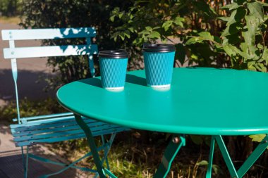 İki kullan-at kahve fincanı, parlak güneş ışığı altında çekilen, arka planda yeşil renkli, uyumlu mavi metal bir sandalyenin yanında canlı yeşil bir masada oturuyor..