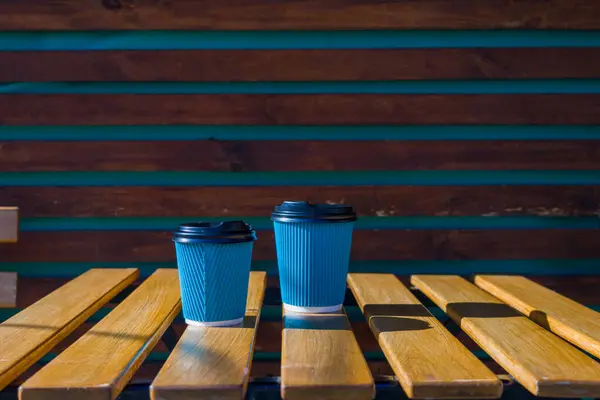 İki adet tek kullanımlık kahve fincanı, turkuaz aksanlı ve doğal güneş ışığıyla sakin bir açık hava ortamında, sade bir ahşap masada oturur. Sıcak atmosferi yükseltir.