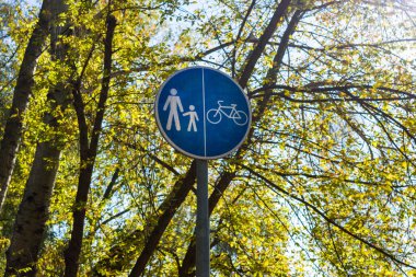 Mavi bir yayanın ve bisiklet yolu tabelasının yakın çekimi. Güneşli bir sonbahar ağacı zeminine karşı. Altından yapraklar yumuşak gölgeler bırakıyor.