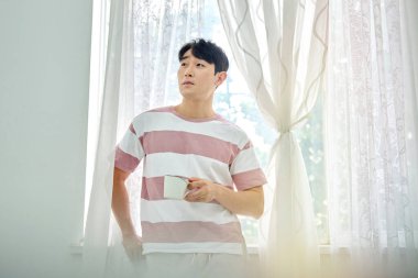 Genç Asyalı Adam Parlak Modern Oturma Odasında Pencere kenarında Kahve İçiyor