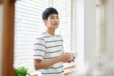 Genç Asyalı Adam Parlak Modern Oturma Odasında Pencere kenarında Kahve İçiyor