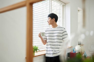 Genç Asyalı Adam Parlak Modern Oturma Odasında Pencere kenarında Kahve İçiyor