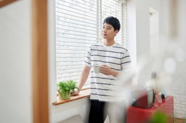 Genç Asyalı Adam Parlak Modern Oturma Odasında Pencere kenarında Kahve İçiyor