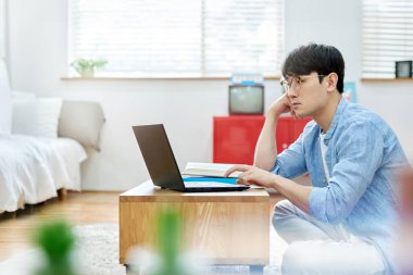 Genç Asyalı Adam Parlak Modern Oturma Odasında Laptop ve Kitapla Çalışıyor