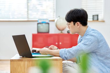 Genç Asyalı Adam Parlak Modern Oturma Odasında Laptop ve Kitapla Çalışıyor